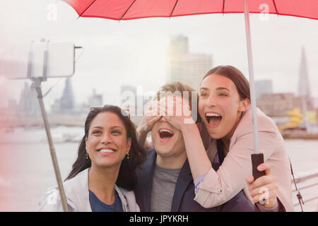 Playful friend tourists with umbrella taking selfie with camera phone selfie stick on bridge, London, UK Stock Photo