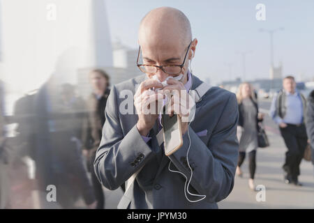 Businessman blowing nose with tissue, holding cell phone and headphones on urban pedestrian bridge Stock Photo