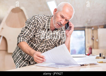 Senior carpenter talking on cell phone and reviewing paperwork in workshop Stock Photo