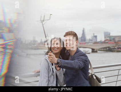 Couple tourists taking selfie with camera phone selfie stick at Thames River waterfront, London, UK Stock Photo