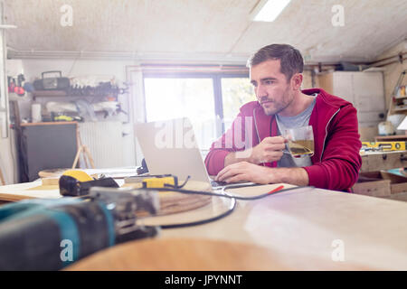 Male carpenter drinking tea and working at laptop on workbench in workshop Stock Photo