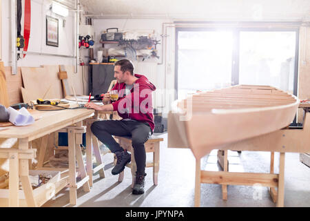 Male carpenter drinking tea and working at laptop on workbench in workshop Stock Photo