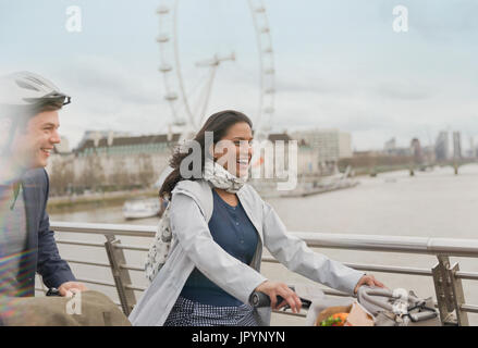 Smiling, carefree couple bike riding on bridge near Millennium Wheel, London, UK Stock Photo