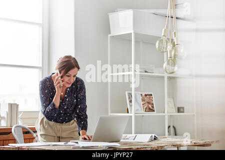 Businesswoman talking on cell phone and working at laptop in office Stock Photo