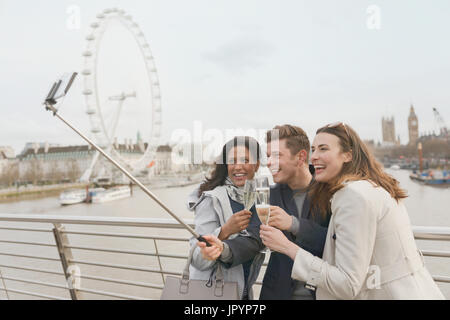 Smiling friend tourists celebrating, toasting champagne and taking selfie with selfie stick near Millennium Wheel, London, UK Stock Photo