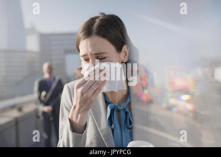 Businesswoman with allergies blowing nose into tissue on sunny urban sidewalk Stock Photo
