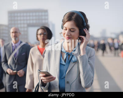 Businesswoman smiling, listening to music with headphones and smart phone on urban pedestrian bridge Stock Photo
