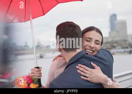 Smiling, affectionate couple with umbrella and flowers hugging on urban bridge, London, UK Stock Photo