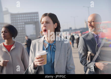 Tired businesswoman with coffee yawning on sunny morning urban pedestrian bridge Stock Photo