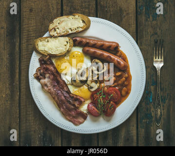 Flat-lay of traditional English breakfast with fried eggs, sausages, mushrooms, sauted tomatoes, bacon, beans and butter toasts on rustic wooden backg Stock Photo