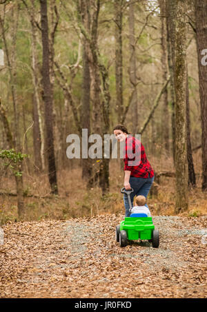 A Caucasian Mother Wearing A Black And Plaid Shirt Pulls Her One-Year Old Son In A Green Wagon On A Gravel Road Covered With Dead Leaves Stock Photo
