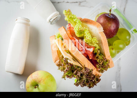 Back to school. A healthy lunch in a box is fresh fruit (apples, plums, grapes), a bottle of yogurt and a sandwich with lettuce, tomatoes, cheese, mea Stock Photo