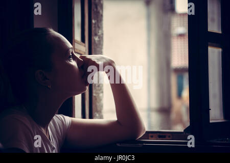 Pensive thoughtful young woman lost in thoughts dreaming and looking in opened  window in vintage style with dramatic mood Stock Photo