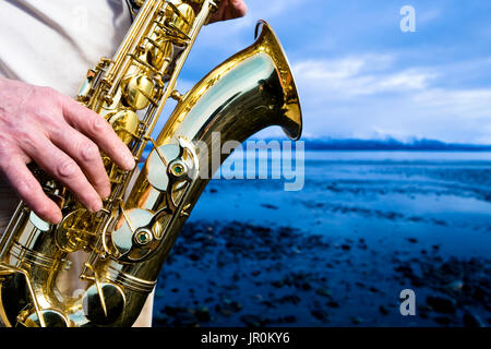 A Man Stands Playing A Saxophone On The Beach At The Water's Edge; Alaska, United States Of America Stock Photo