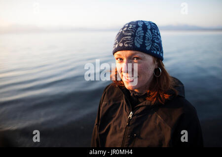 Portrait Of A Woman With Red Hair Wearing A Knit Hat And Standing With Water In The Background; Alaska, United States Of America Stock Photo