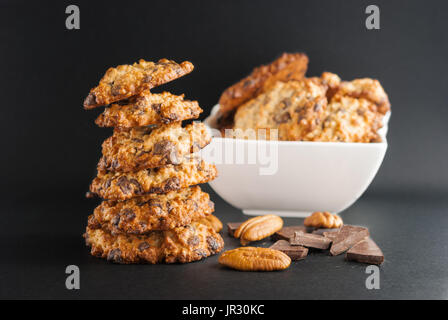 Stacked chocolate chip cookies, white bowl with cookies, pecan and pieces of chocolate on a black background Stock Photo