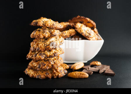 Stacked chocolate chip cookies, white bowl with cookies, pecan and pieces of chocolate on a black background Stock Photo