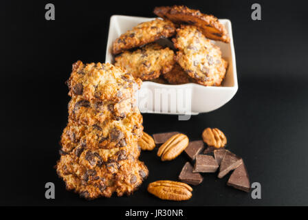 Stacked chocolate chip cookies, white bowl with cookies, pecan and pieces of chocolate on a black background Stock Photo