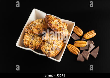 Chocolate chip cookies in a white bowl, pecan nuts and pieces of chocolate, top view,  on a black background Stock Photo