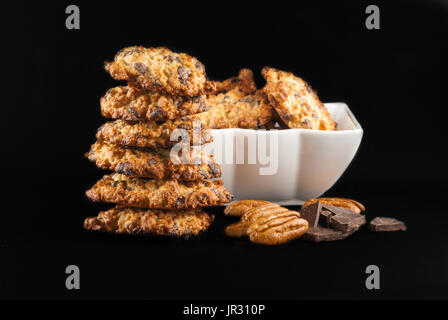 Stacked chocolate chip cookies, white bowl with cookies, pecan and pieces of chocolate on a black background Stock Photo