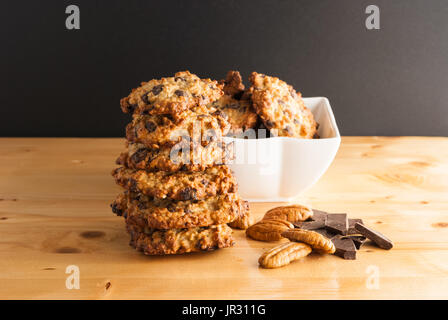 Stacked chocolate chip cookies, white bowl with cookies, pecan and pieces of chocolate on a black background Stock Photo