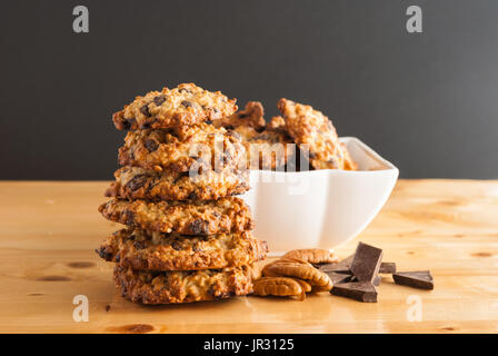 Stacked chocolate chip cookies, white bowl with cookies, pecan and pieces of chocolate on a black background Stock Photo