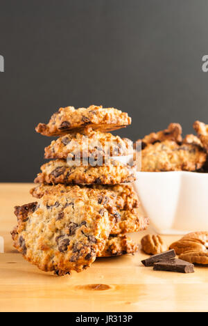 Stacked chocolate chip cookies, white bowl with cookies, pecan and pieces of chocolate on a black background Stock Photo