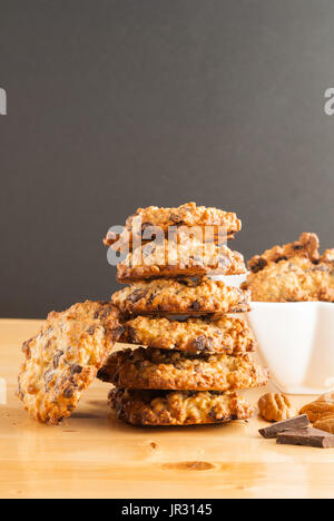 Stacked chocolate chip cookies, white bowl with cookies, pecan and pieces of chocolate on a black background Stock Photo