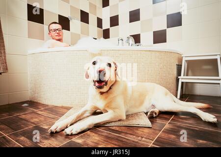 Living with dog. Young man relaxing in bathroom with his yellow labrador retriever. Stock Photo