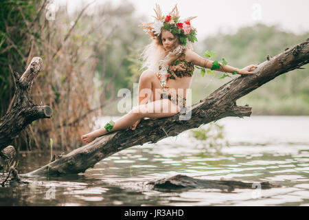 Beautiful little girl in image of nymph with floral head wreath sits on tree slanted above water. Stock Photo