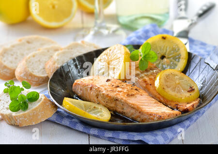 Two fried salmon fillets with steamed slices of lemon in a pan,  served with baguette, white wine in the background Stock Photo
