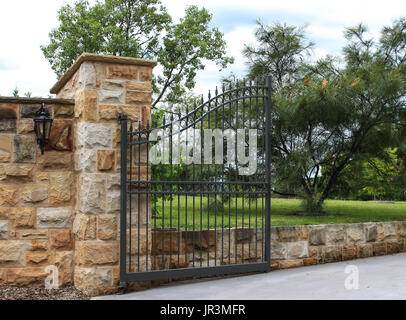 Black metal driveway entrance gate set in sandstone brick fence with garden trees in background Stock Photo