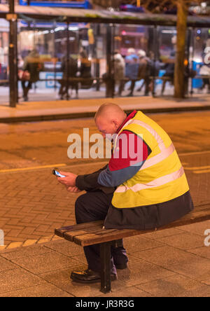 Bus driver  wearing a uniform sitting on a bench reading his mobile phone on the street at night in Canterbury, Kent. Stock Photo