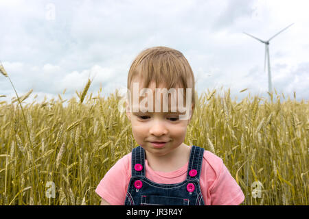 little girl child is standing in crop wheat field in front of wind turbine plant farm Stock Photo