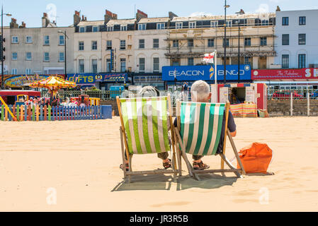 A mature couple sitting together on stripy deckchairs on the sandy beach at Margate, Kent, facing the buildings on the seafront. The weather is warm. Stock Photo