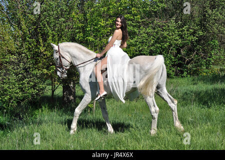 young attractive woman in white dress horseback riding on white horse in the summer meadow Stock Photo