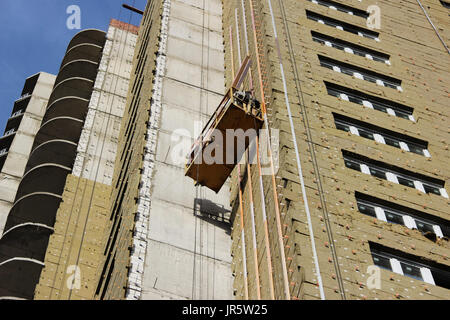 worker in yellow suspended cradle mounts environmental boards for insulation on a newly built high-rise building. Stock Photo