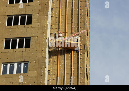 worker in yellow suspended cradle mounts environmental boards for insulation on a newly built high-rise building. Stock Photo