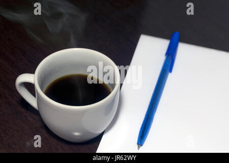 White porcelain smoking and steaming hot coffee mug, blank notebook and blue pen on top of a dark wood table Stock Photo