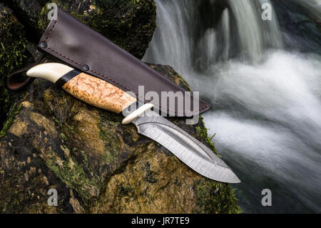 Damascus hunting knife with scabbard on the stone by the river. Stock Photo