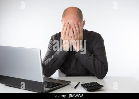 Despairing hopeless man sitting with his head in his hands as he sits doing his accounts at his laptop computer Stock Photo