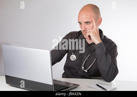Doctor with a stethoscope around his neck sitting working at his laptop computer resting his chin on his hand with a serious thoughtful expression Stock Photo