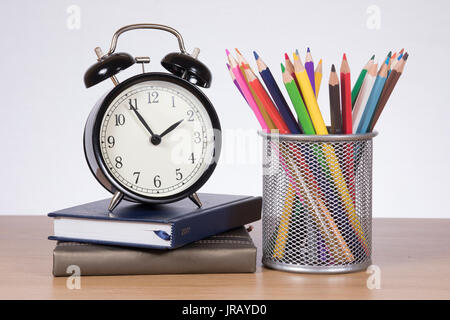 Stacked books with an alarm clock and coloured pencils on a timber desk in a time to learn, back to school theme. Stock Photo