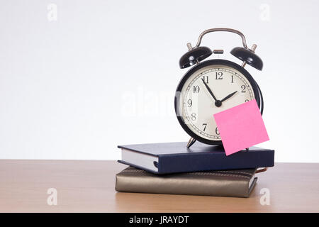 Alarm clock with sticky note standing on pile of calendars on desk against white background Stock Photo