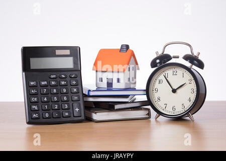Conceptual image with calculator propped up to show the display, model house and alarm clock standing on a pile of books on a wooden table Stock Photo