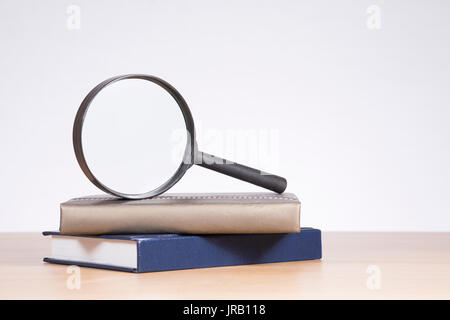 Magnifying glass balanced on top of two hardcover books with copy space over a white wall above a wooden table Stock Photo