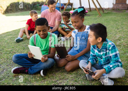 Children using digital tablet while sitting with man on field at park Stock Photo