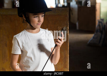 Thoughtful girl holding crop stick in stable Stock Photo