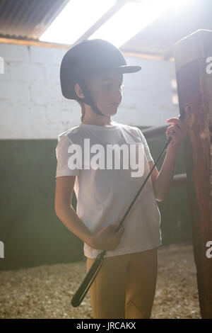 Thoughtful girl holding crop stick in stable Stock Photo