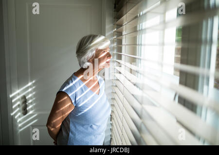 Senior woman looking through window in bedroom at home Stock Photo